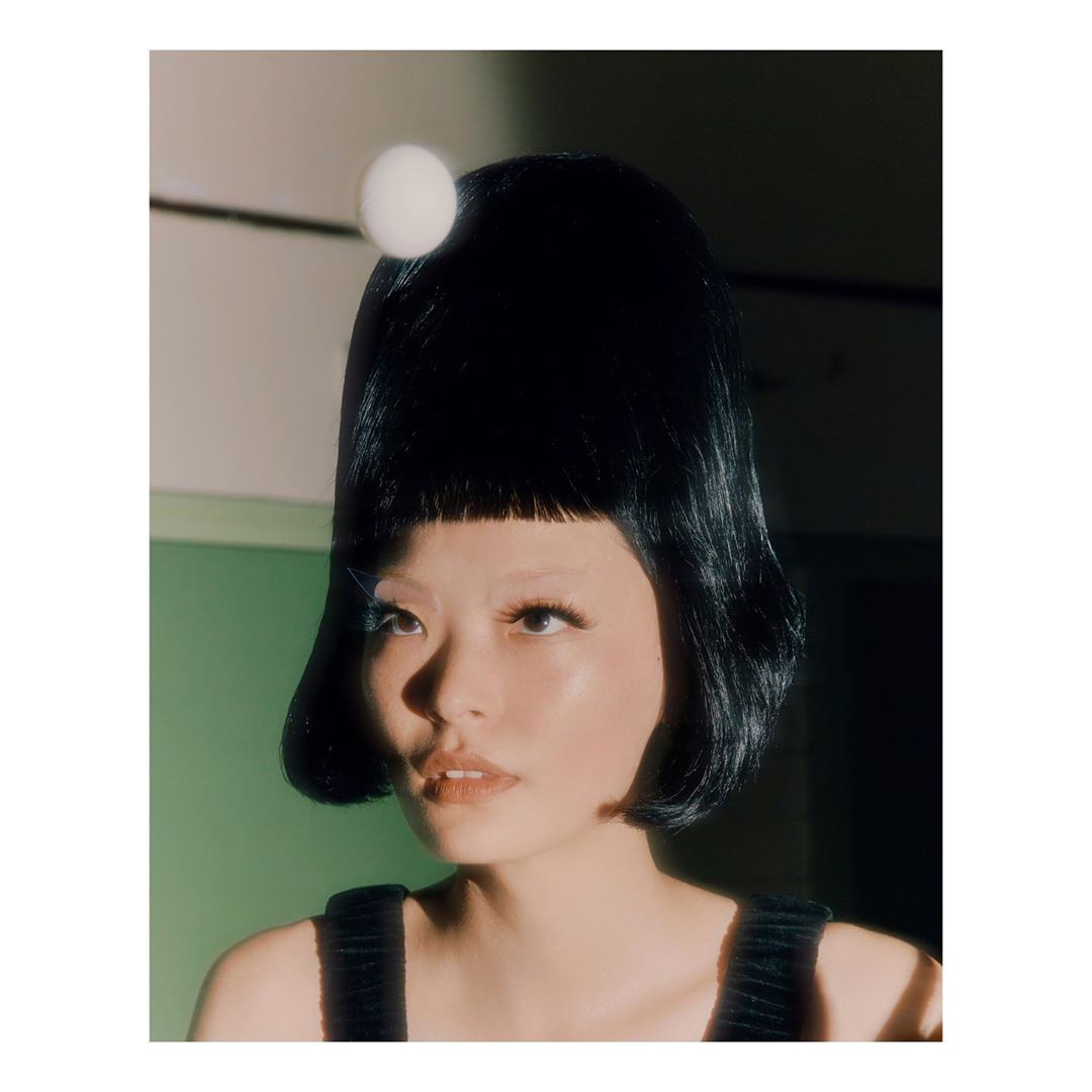 Close-up of a stylish woman with a black bob haircut and a white ball on her head, wearing a black top, against a green background.