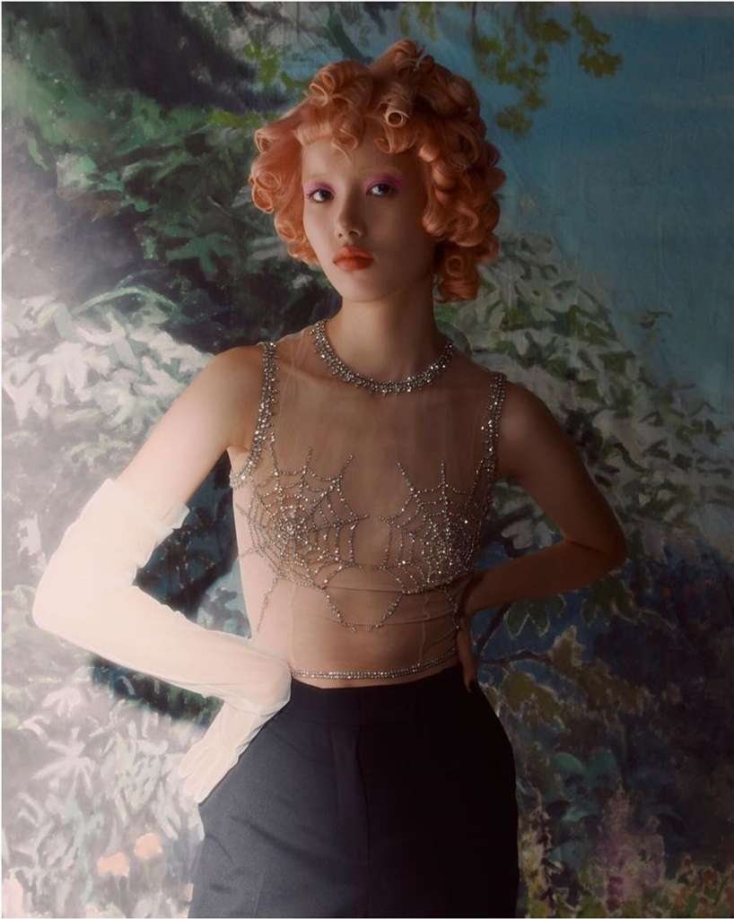 Portrait of a stylish Asian woman with orange curly hair and a beaded top, set against a dreamy, artistic floral backdrop.