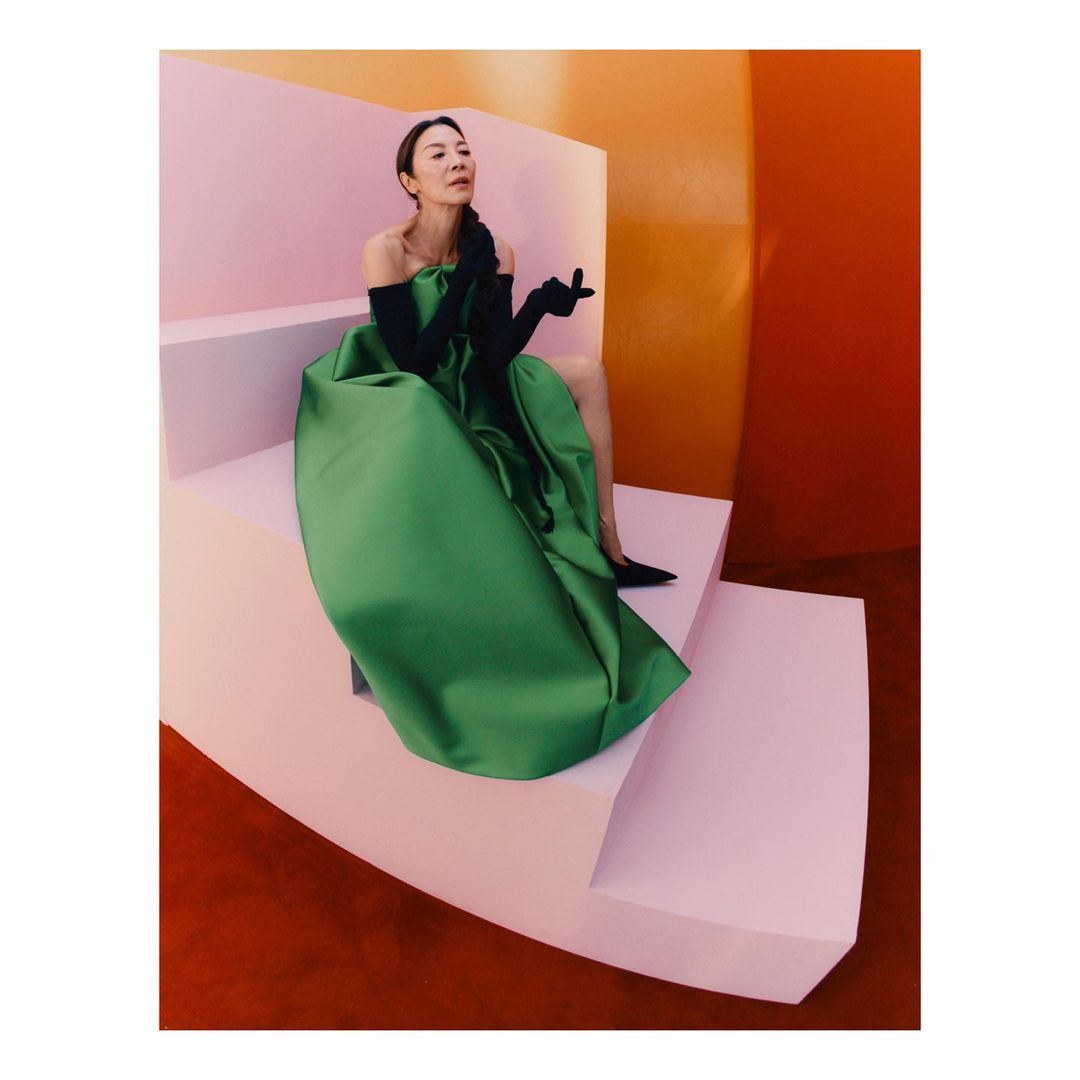 Michelle Yeoh sits on pink steps in a voluminous green gown with black gloves against an orange backdrop, a high-fashion portrait.