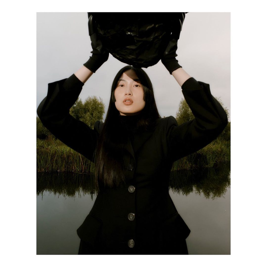 A woman in a black coat holds a dark, abstract object above her head against an outdoor backdrop of greenery and water reflections.