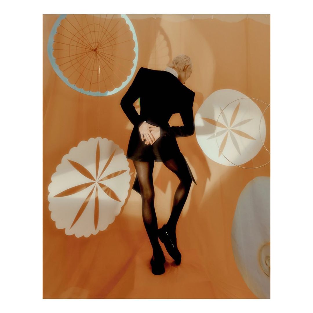 Fashionable adult with blonde hair poses in a black jacket and tights in an indoor photo studio with peach-colored walls.
