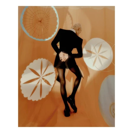 Fashionable adult with blonde hair poses in a black jacket and tights in an indoor photo studio with peach-colored walls.