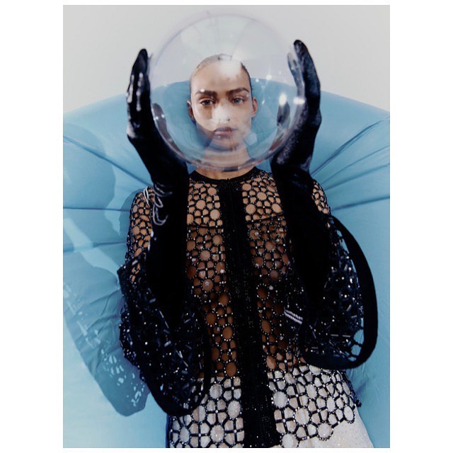 Woman in sheer beaded dress, black gloves, and clear space helmet posing on turquoise beanbag chair against a white background.