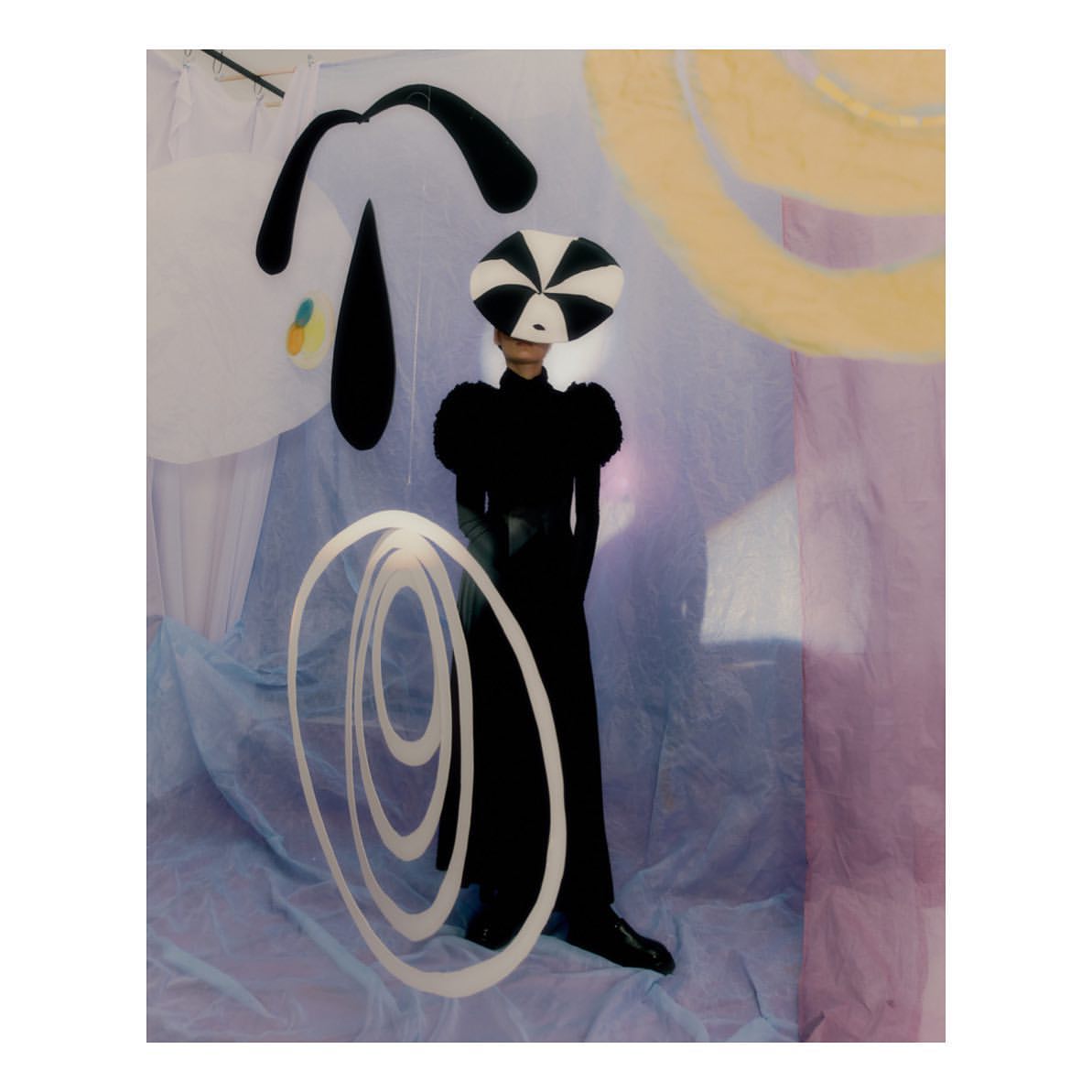 A person wearing a black dress and a black and white hat poses in front of abstract black and white paintings with a light purple backdrop.
