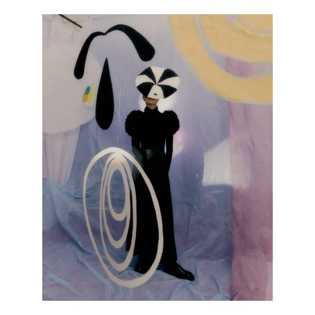 A person wearing a black dress and a black and white hat poses in front of abstract black and white paintings with a light purple backdrop.