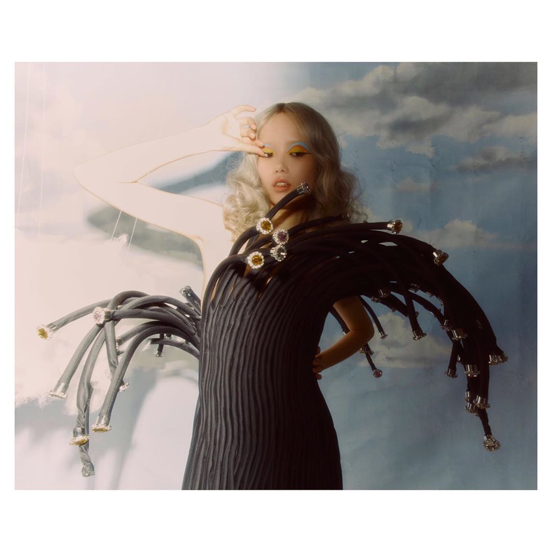 Editorial fashion shot: a young woman with blond curly hair poses in a black dress with elaborate tubing against a cloudy backdrop.