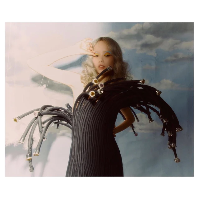 Editorial fashion shot: a young woman with blond curly hair poses in a black dress with elaborate tubing against a cloudy backdrop.
