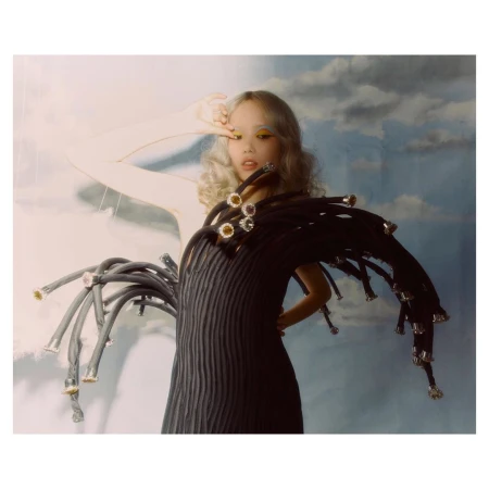 Editorial fashion shot: a young woman with blond curly hair poses in a black dress with elaborate tubing against a cloudy backdrop.