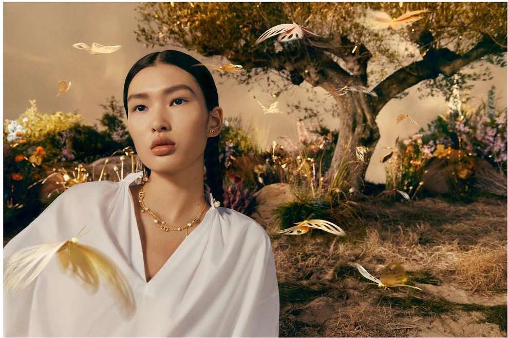 Woman in white blouse and gold necklace poses near a tree in a surreal, dreamlike scene with decorative golden butterflies.
