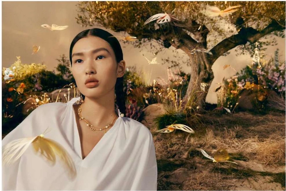 Woman in white blouse and gold necklace poses near a tree in a surreal, dreamlike scene with decorative golden butterflies.