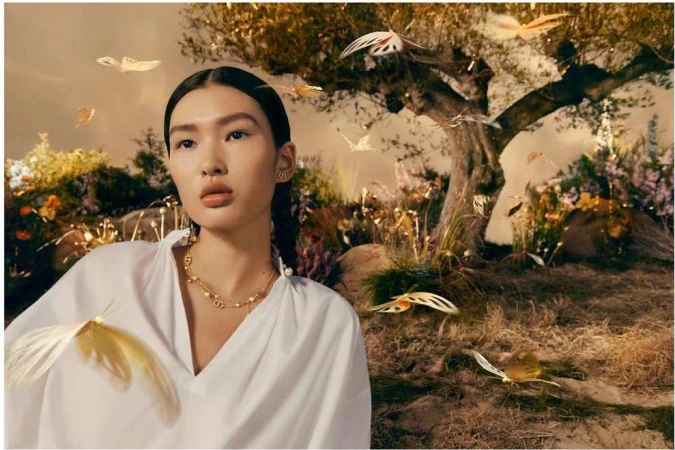 Woman in white blouse and gold necklace poses near a tree in a surreal, dreamlike scene with decorative golden butterflies.