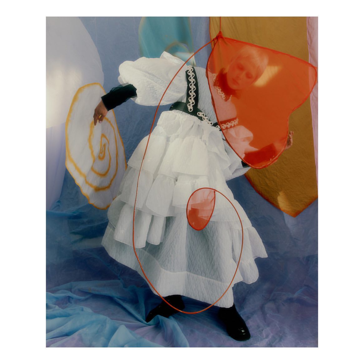 A person poses in a white tiered dress with black trim and shoes, accented by orange shapes, against a multicolored background.