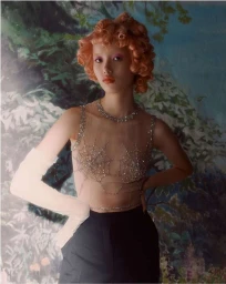 Model with orange curly hair wearing a sheer crystal-embellished top with a spiderweb design and dark trousers.