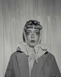 Portrait of a woman wearing a polka dot headscarf and a blazer against a draped curtain background.