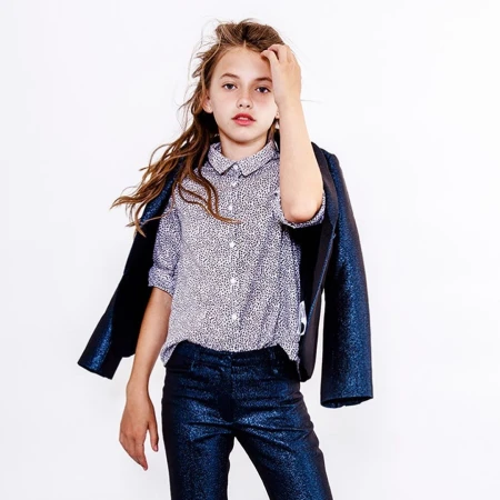 Young girl with long brown hair wearing a patterned button-down shirt, dark trousers, and a dark jacket draped over her shoulders.