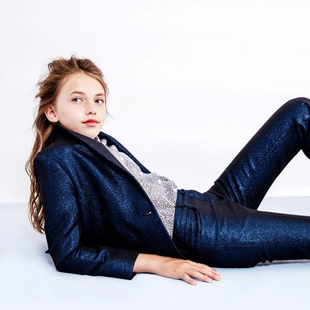 A young girl in a sparkly blue pantsuit lies at an angle against a white backdrop looking off-camera, portraying a fashion editorial.