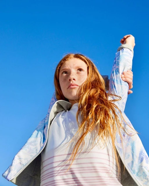 Young girl with red hair in casual clothing looking up at a blue sky with her arm raised overhead in an outdoor setting.