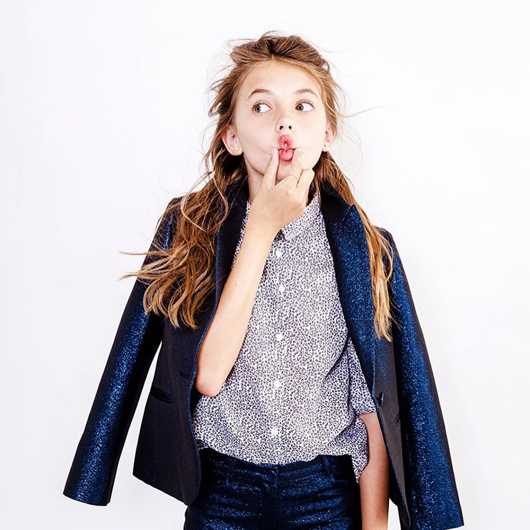 Girl in a shiny blue blazer and spotted shirt poses playfully against a white backdrop, looking off-camera with her finger near her lips.