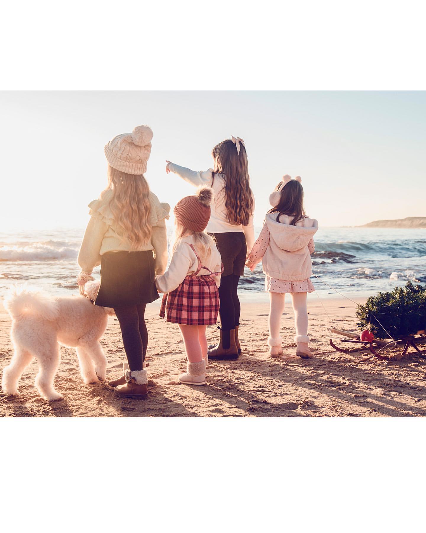 Four young girls and a dog stand on a sandy beach near the ocean waves with a small Christmas tree on a sled in golden sunlight.