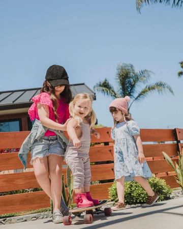 Three young children playing with a skateboard outdoors near a wooden fence under a clear sky.