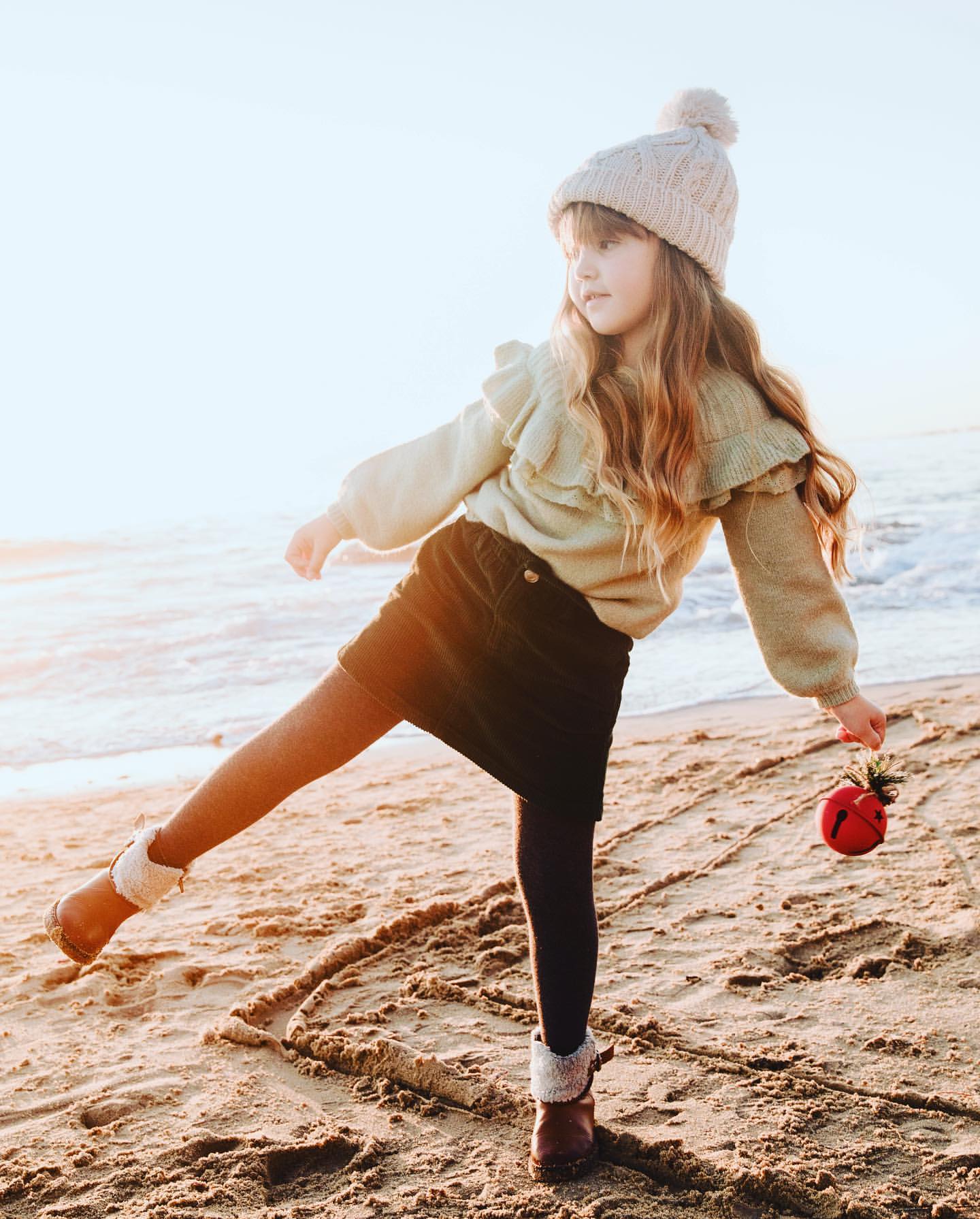 A young girl in a knit hat and sweater poses on a beach at sunset, holding a red ornament and drawing in the sand with her foot.