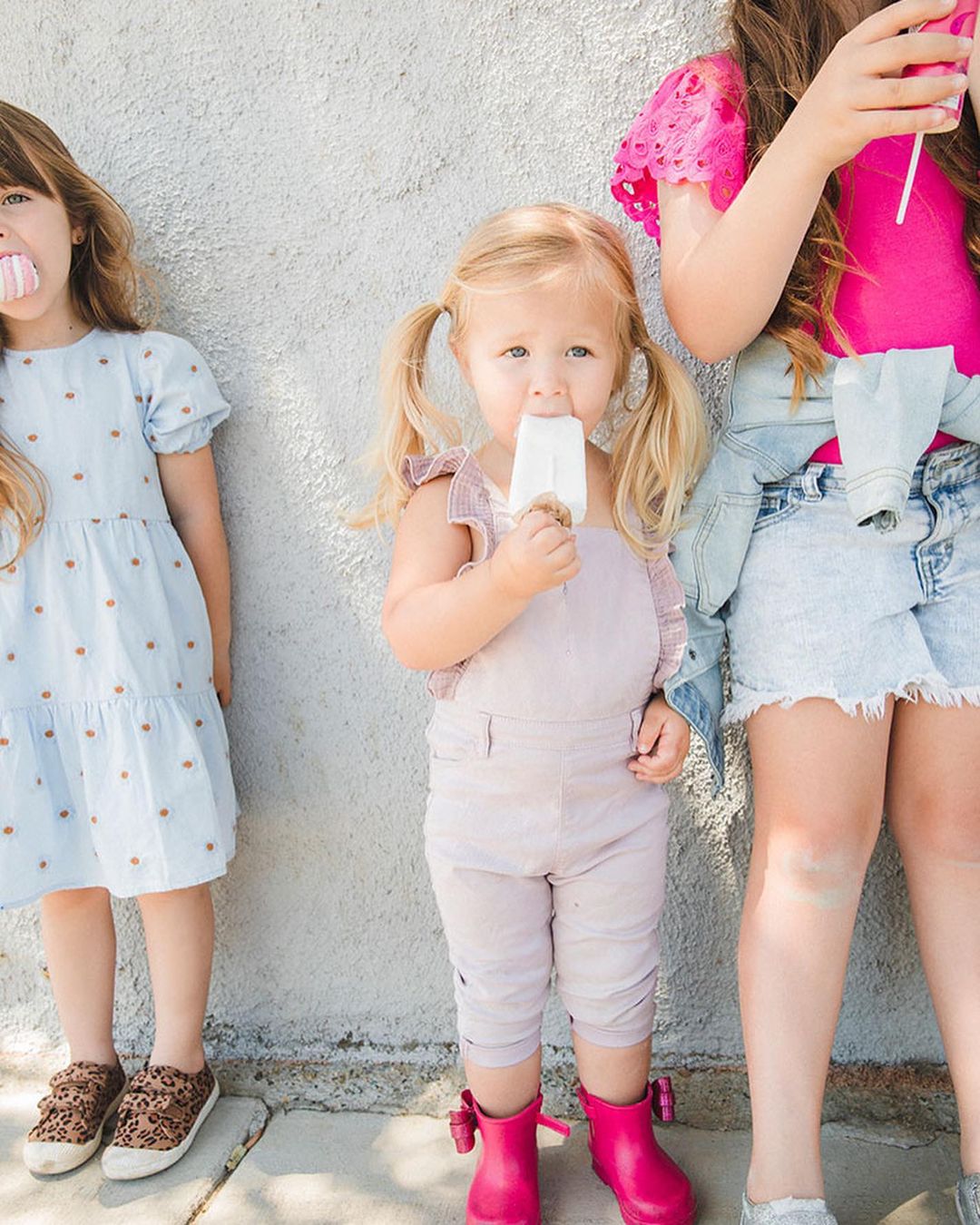 Three young girls stand outside a building while eating popsicles; one girl wears pink boots and overalls, and another has a blue dress.