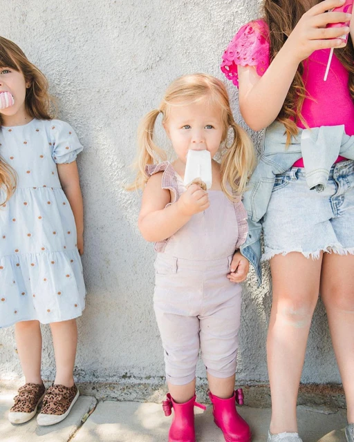 Three young girls stand outside a building while eating popsicles; one girl wears pink boots and overalls, and another has a blue dress.