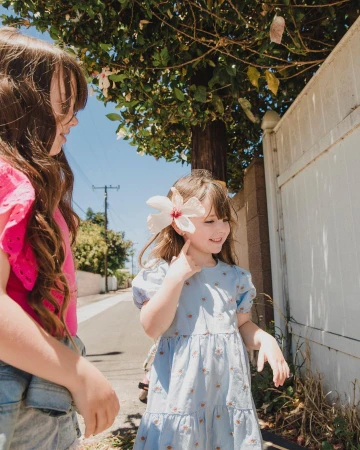 Two young girls standing outdoors on a sunny street, one wearing a blue dress with a large white flower behind her ear