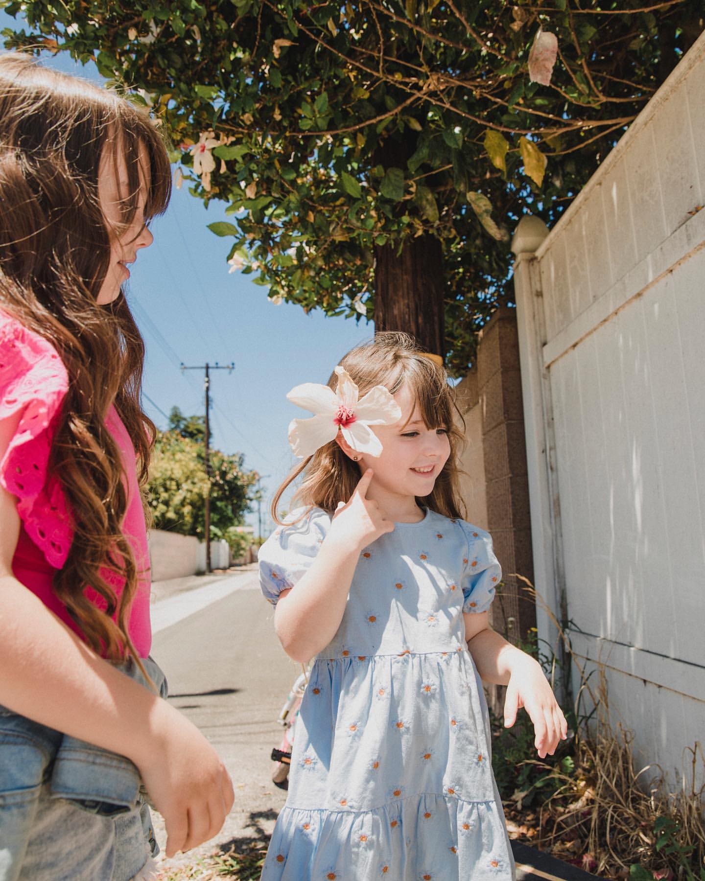 Two young girls outdoors with a large white flower tucked behind one child's ear, standing near a white fence and trees.