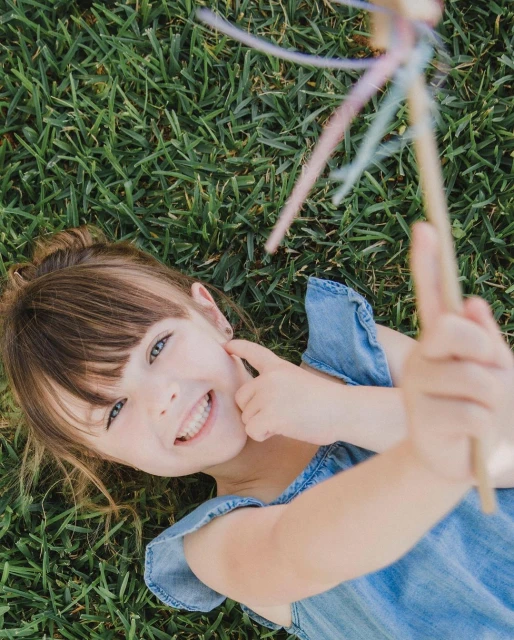 Young girl lying in green grass smiles, holding a wand with ribbons in an eye-level portrait with shallow depth of field on a sunny day.