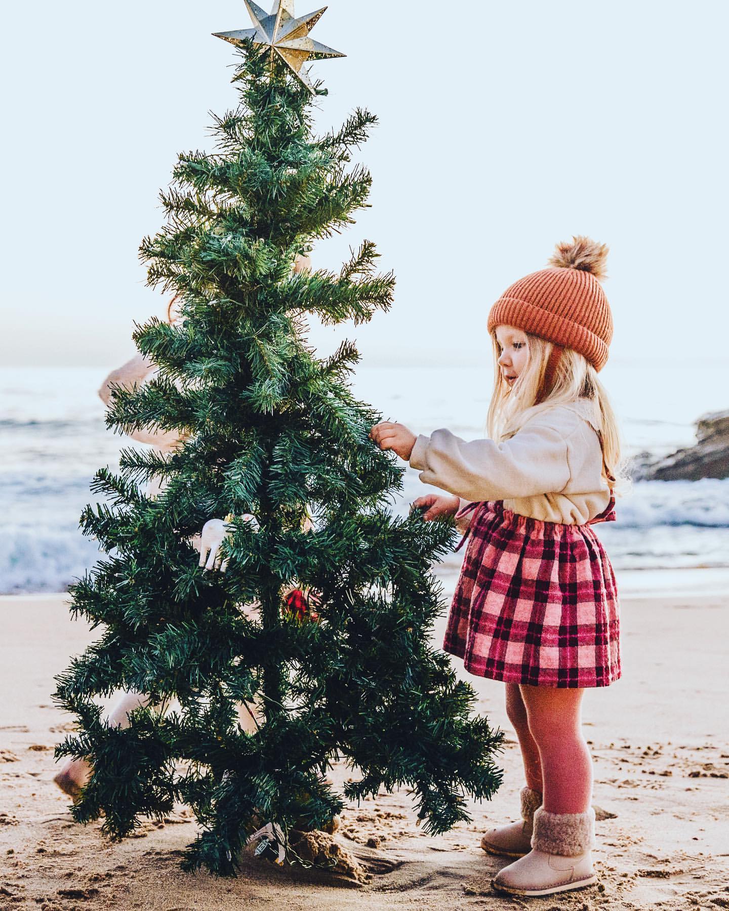 Adorable child decorating a small Christmas tree on a sandy beach at daytime, wearing winter clothing and a beanie; bright and festive.