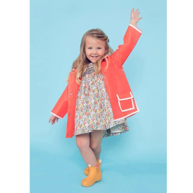 A young girl smiles while wearing a floral dress, coral jacket with white trim, and yellow rain boots against a blue background.