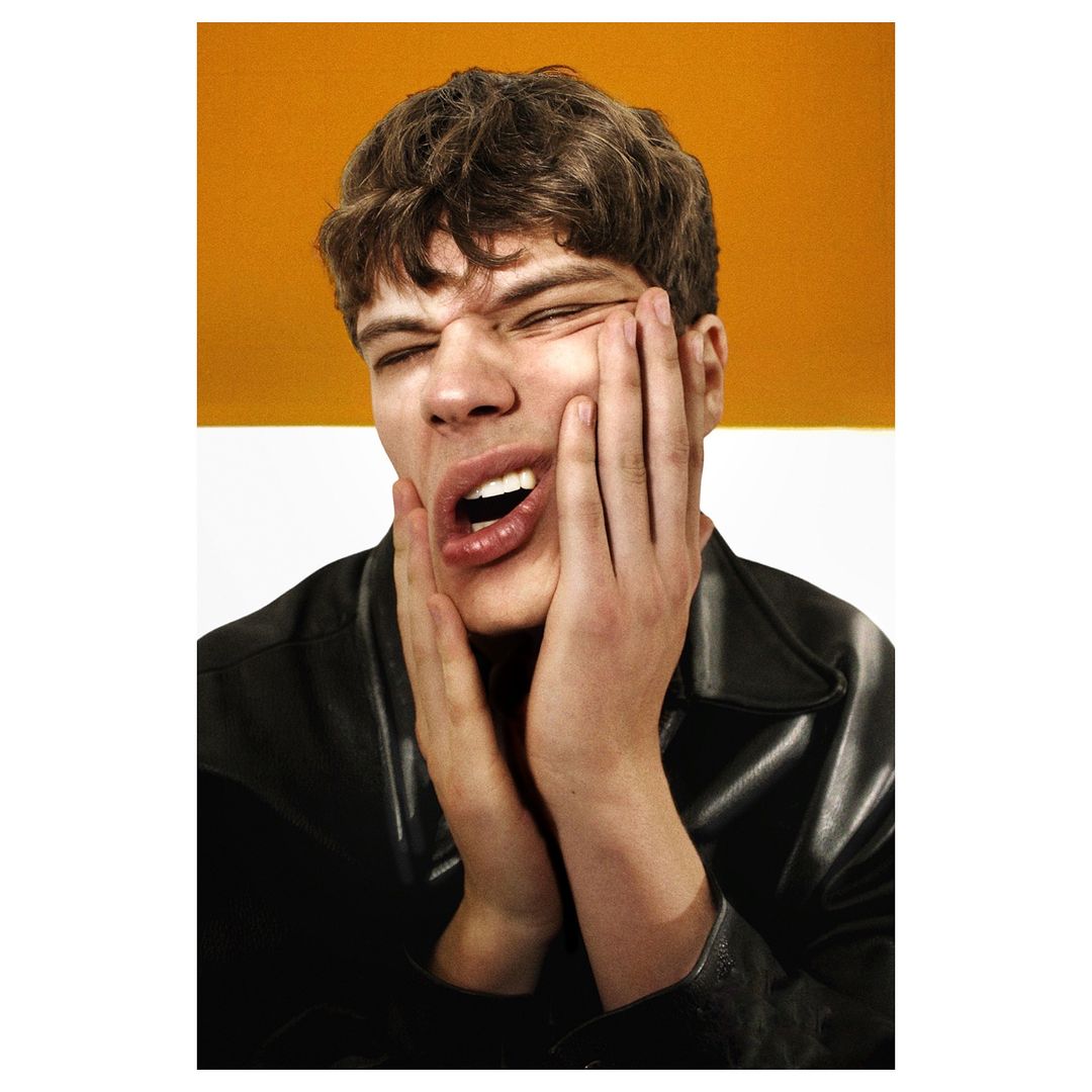 Close-up portrait of a young adult man with dark hair, mouth open, and hands on his face, against a split orange and white background.