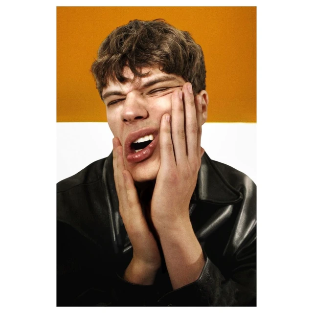 Close-up portrait of a young adult man with dark hair, mouth open, and hands on his face, against a split orange and white background.