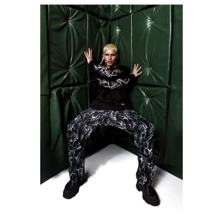 Adult poses in black printed sweater and matching pants against a green padded backdrop in an indoor studio setting.