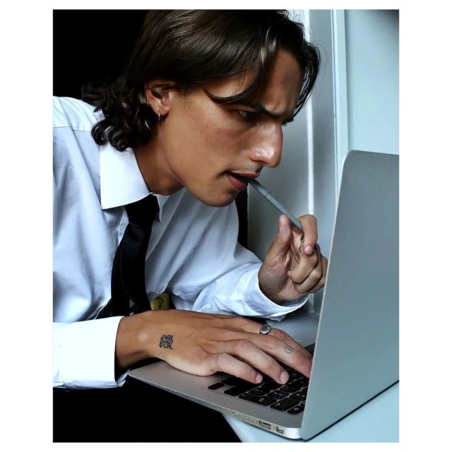 Thoughtful adult male in a white shirt with a pen in his mouth works at a laptop with a neutral background in a close-up shot.