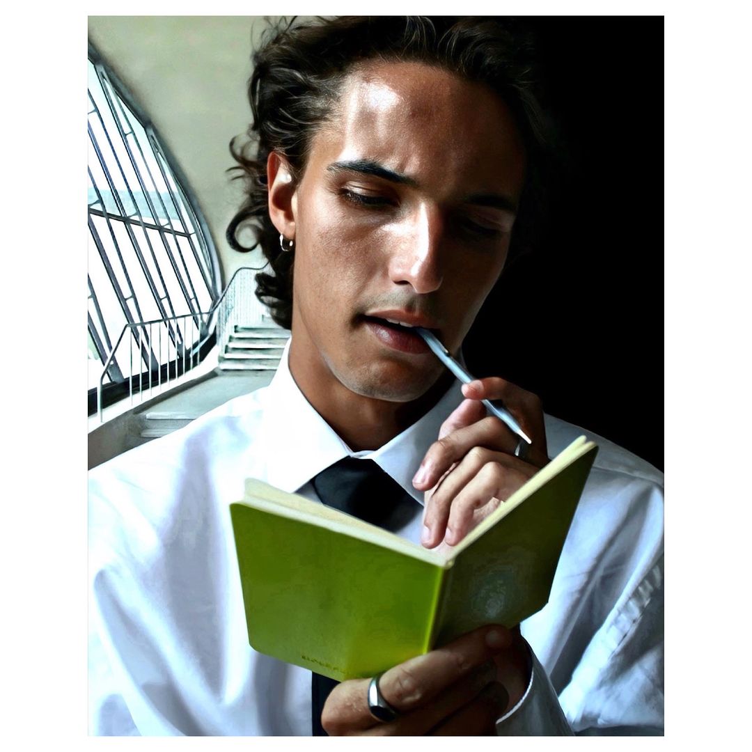 Young adult male in white shirt and black tie holding pen and open green notebook indoors with modern staircase background.