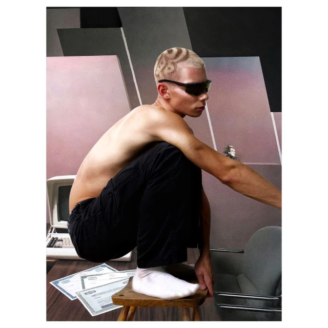 A shirtless man with patterned buzzcut sits on a wooden stool wearing sunglasses, with computer and documents surrounding him in studio.