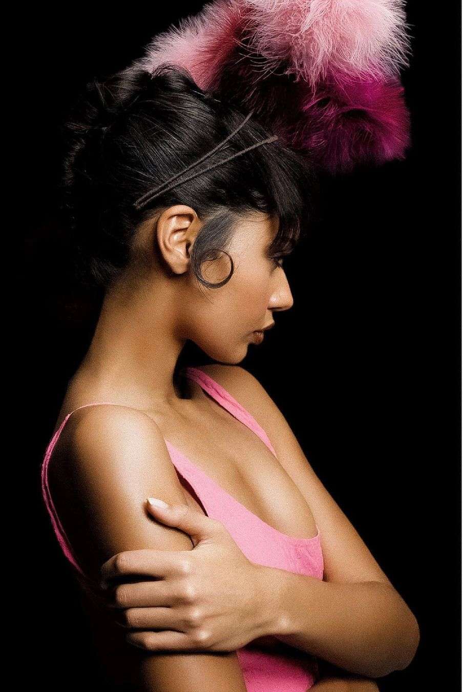 Profile of a woman with dark hair, wearing a pink tank top, and a headpiece with feathers against a dark background.