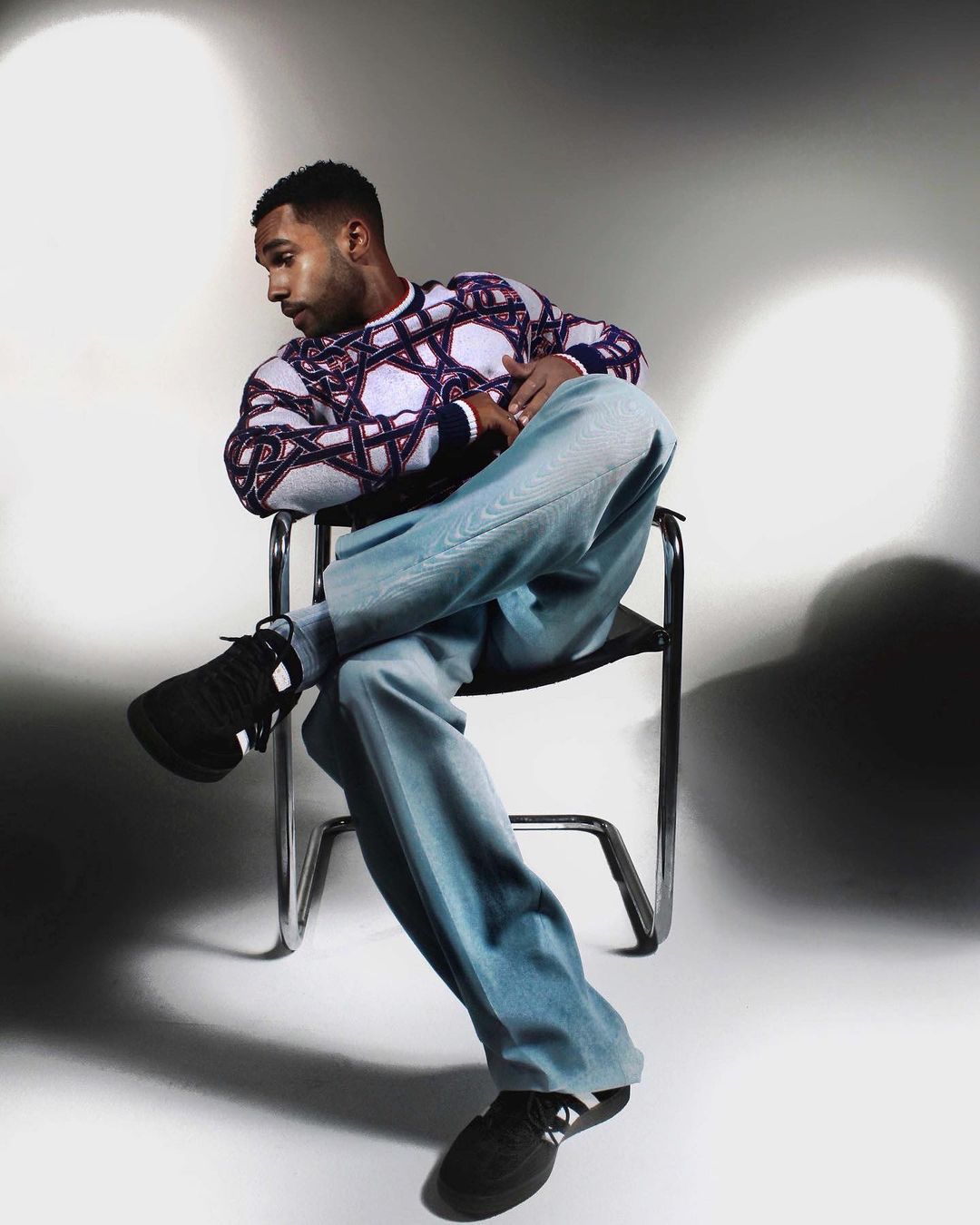 Portrait of an adult male in a patterned sweater sitting on a chrome chair in a photo studio with soft lighting. He is looking away.