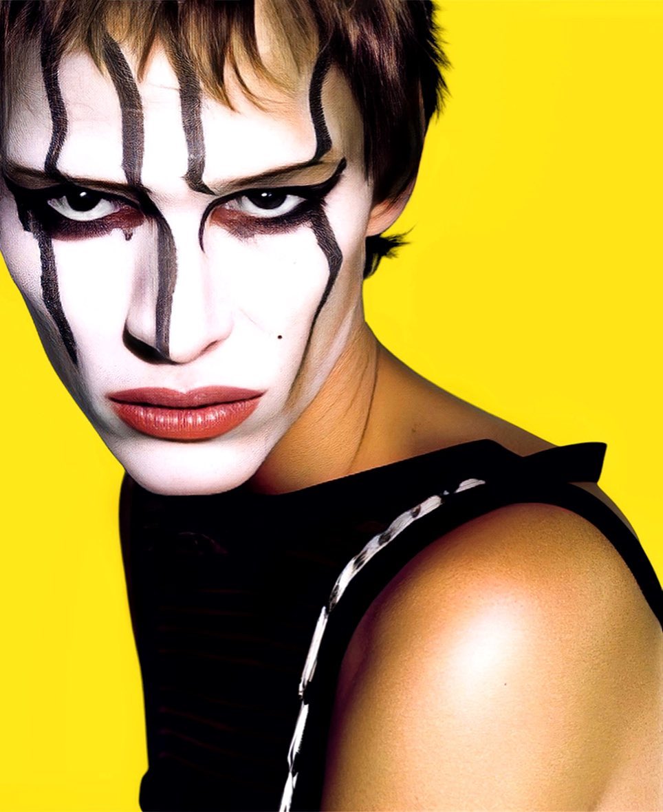 Close-up portrait of a person with dramatic black and white face makeup, short brown hair, black top, on a vivid yellow background.