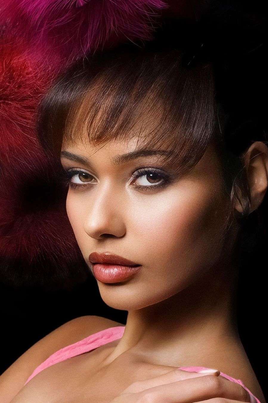 Close-up portrait of a woman with brown hair and neutral makeup, adorned with a red feathered accessory against a black background.