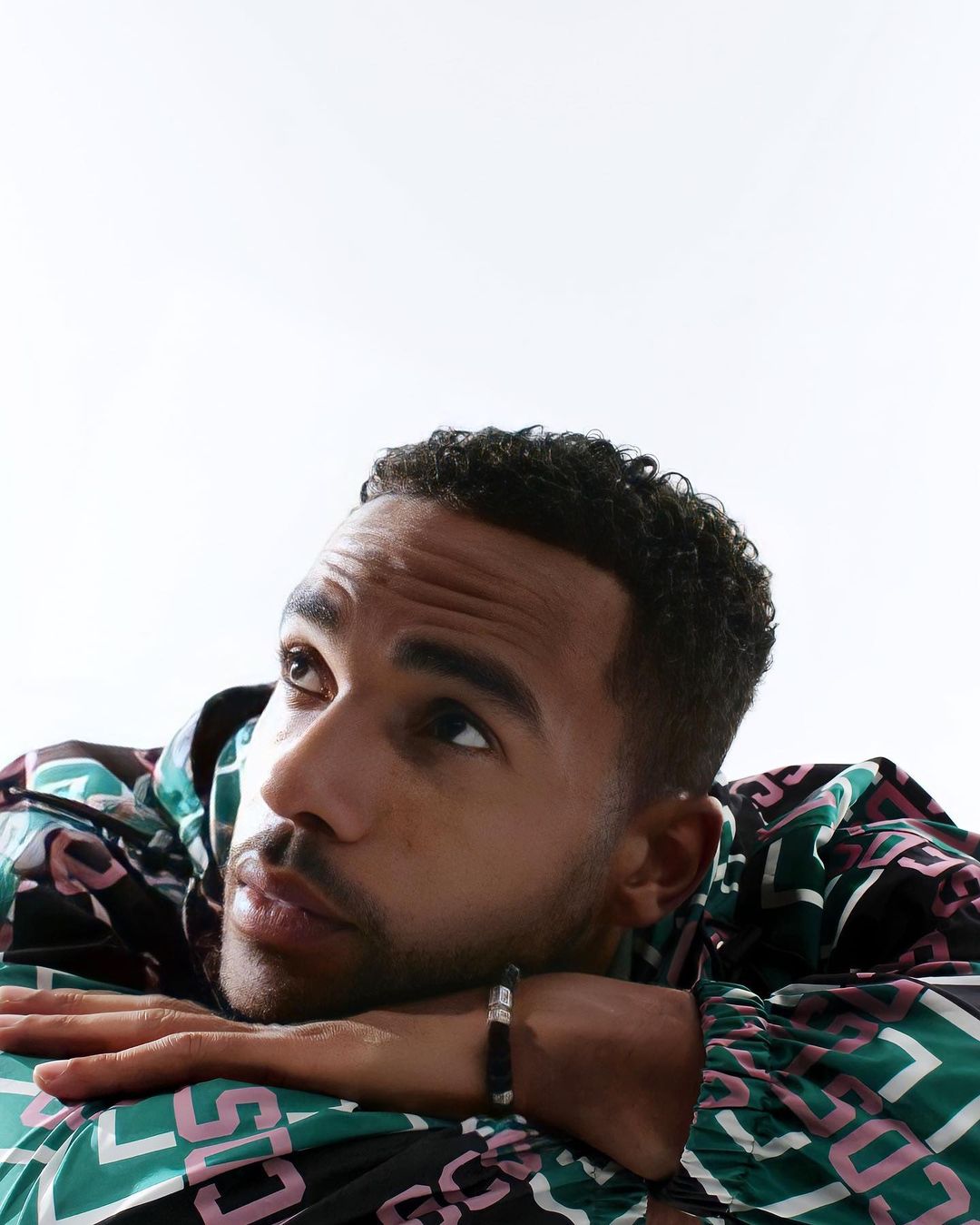 Portrait of a young adult male resting his chin on his hands, gazing upward against a bright white background, wearing a patterned jacket.