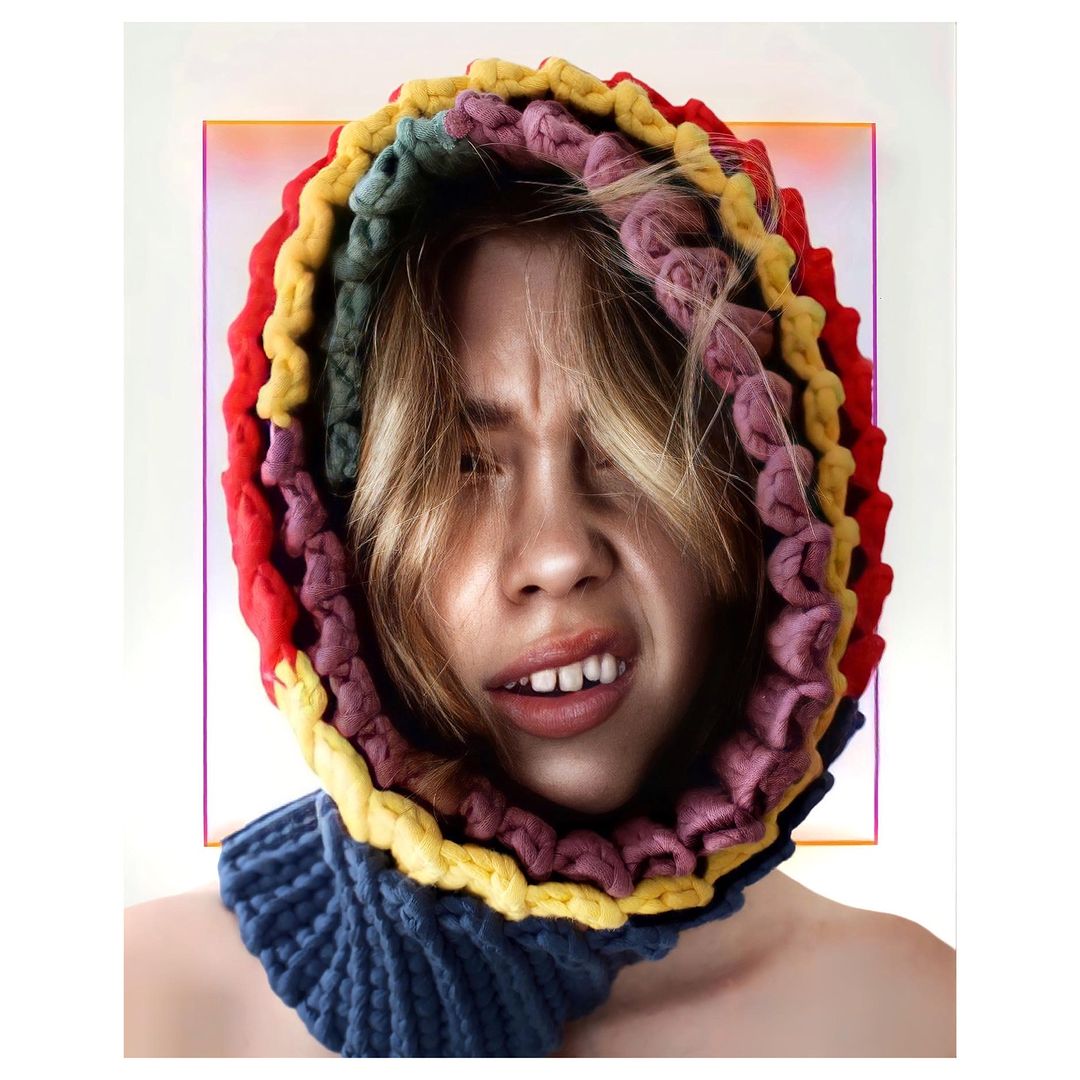 Close-up portrait of a woman with a scrunched face and a rainbow knitted hood and collar, set against a clean white background with a colored frame.