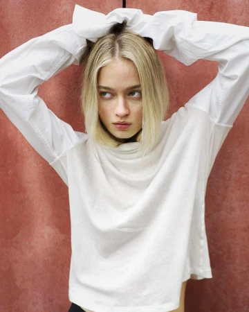 Blonde woman with hands in her hair wearing a white long-sleeve top against a textured red wall background.