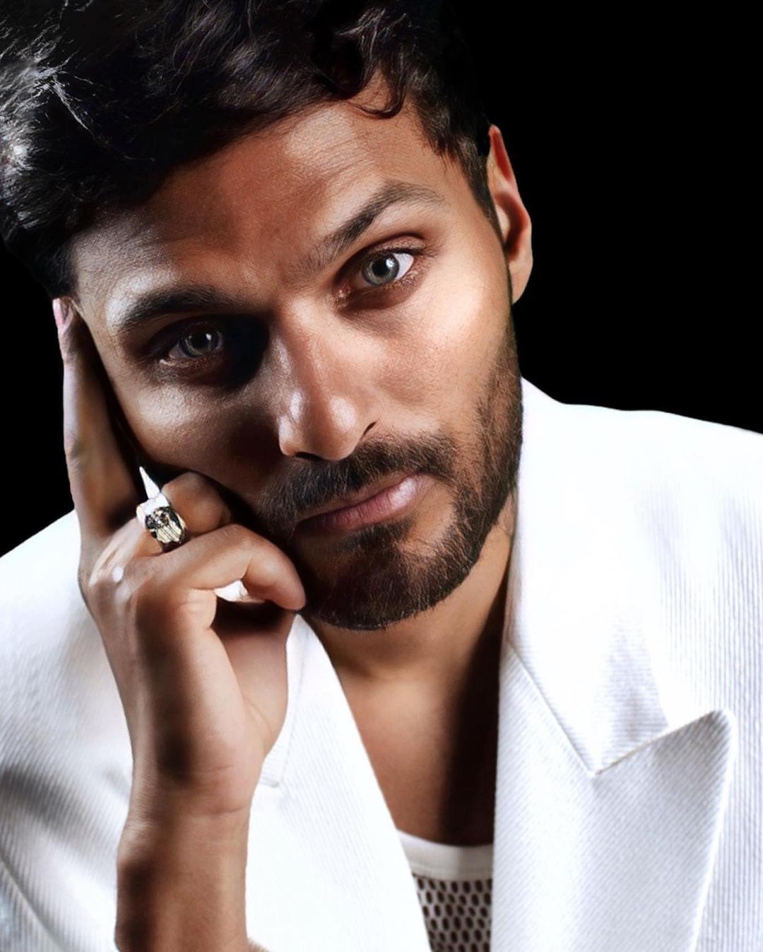 Close-up portrait of a man with a beard wearing a white suit. He is resting his face on his hand with a ring on his finger.