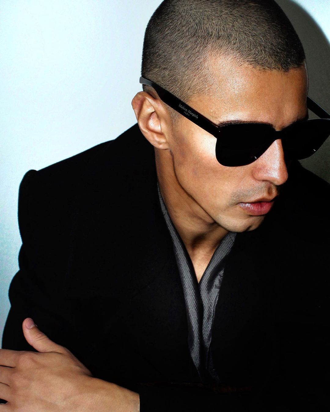 Adult male poses indoors wearing a black coat, gray collared shirt, and sunglasses against a gradient background. High-fashion mood.