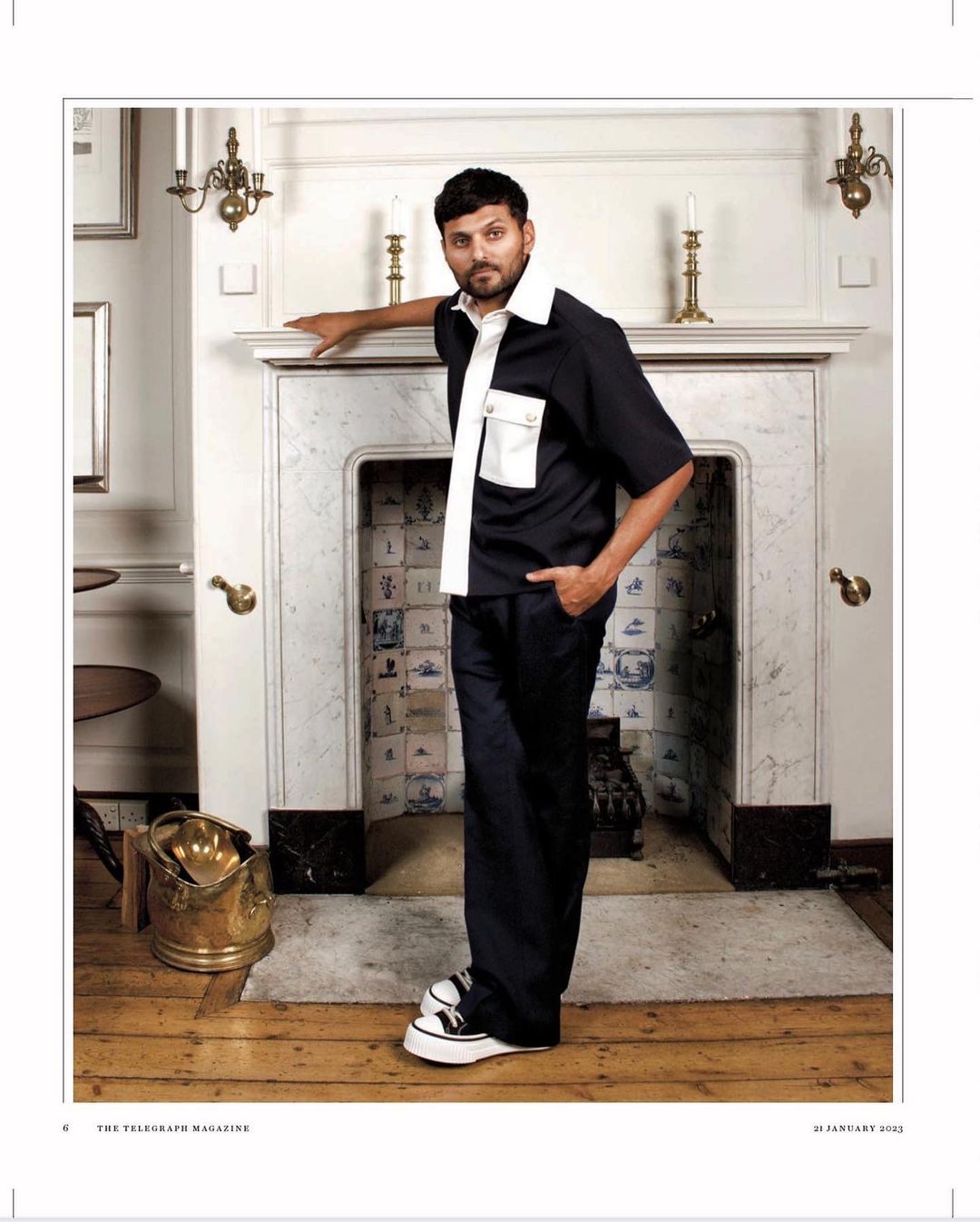 Adult male in a black and white short-sleeved outfit poses next to a vintage fireplace with brass objects on a wood floor indoors.