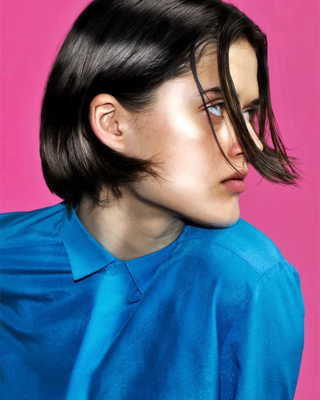 Close-up studio portrait of a young adult woman with short dark hair, wearing a blue shirt, set against a vibrant pink background.