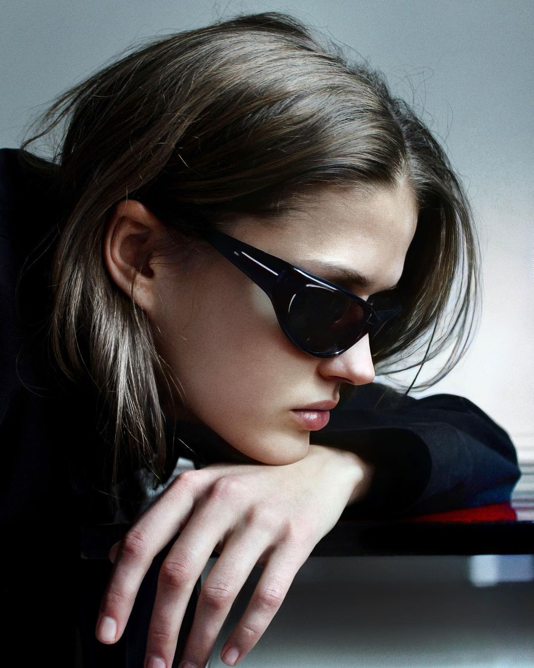 Portrait of a young adult woman wearing black sunglasses, posing indoors with natural light and a minimalist backdrop, fashion editorial.